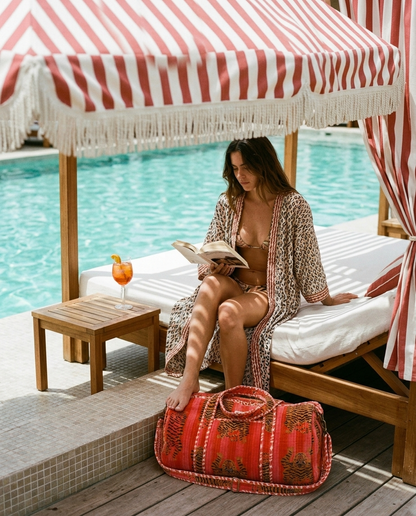Poolside daybed reading under a striped fringed canopy with the Tibetan Tiger Velvet Weekender in Orange and Pink at her feet — handmade in Rajasthan by Conscious Yoga Collective
