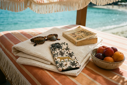 The Jungle iPhone Case in Cream resting on a folded Turkish towel on a striped cabana daybed with a bowl of stone fruit