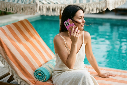 The Tibetan Tiger iPhone Case in Pink held to the ear on a striped cabana lounger beside a Mediterranean pool