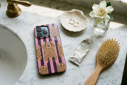 The Tibetan Tiger iPhone Case in Mauve Plum on a white marble bathroom counter with gold hoops and gardenia